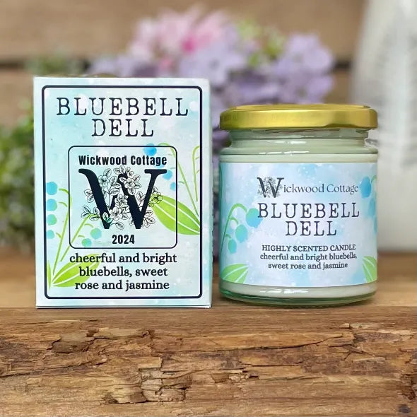Wickwood Cottage Bluebell Dell candle and packaging on a rustic wooden surface with a blurred floral background. To the left is the candle box pale blue with bluebell florals and black text. To the right is the candle a 150-gram candle jar with a gold lid.