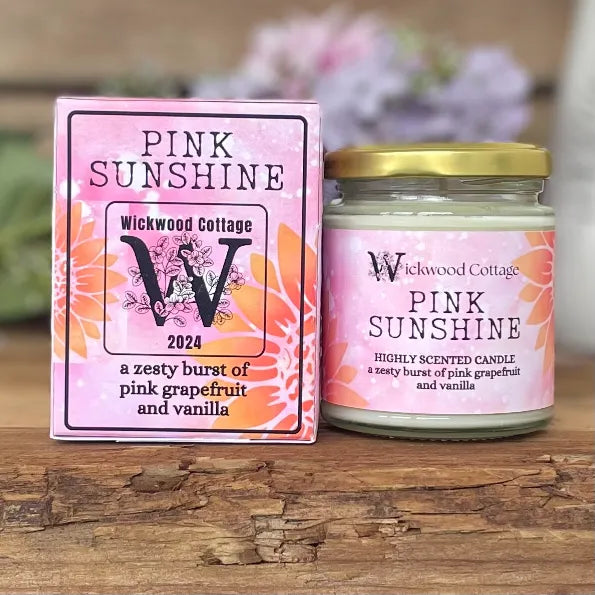 Wickwood Cottage Pink Sunshine candle and packaging on a rustic wooden surface with a blurred floral background. To the left is the candle box pink with a yellow sunflower and black text. To the right is the candle a 150-gram candle jar with a gold lid.