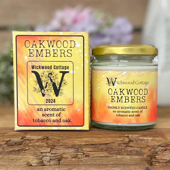 Wickwood Cottage Oakwood Embers candle and packaging on a rustic wooden surface with a blurred floral background. To the left is the candle box yellow and red with black text. To the right is the candle a 150-gram candle jar with a gold lid.
