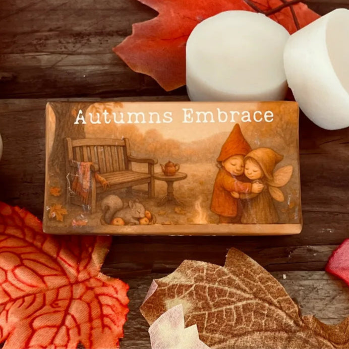 Paper wrapped rectangle wax melt with 'Autumns Embrace' text and an image of an enchanted fae couple hugging with a wooden bench to the left. Surrounding the package, autumn leaves a pumpkin and two wax melt discs on a rustic wooden surface. Close-up image