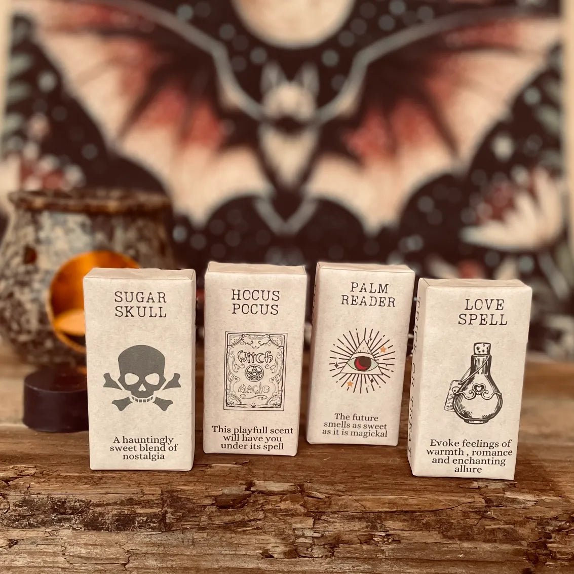 Four wax melts wrapped in vintage paper with spooky label designs and informative text. With a blurred background of a bat and wax warmer on a rustic wooden surface.