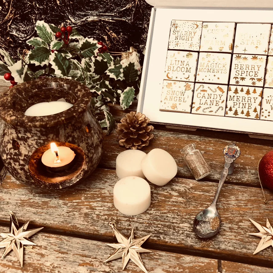 An enchanted image of an open white Christmas box full of square individually wrapped wax melts in 12 different fragrances. Each melt is wrapped in white paper with gold text. The box sits on a rustic wooden surface to the left is a piece of holly and a pinecone. A rustic looking wax warmer with a glowing tealight and melting wax. In the foreground three white wax melt discs a spoon and a small bottle of glitter all resting on a rustic wooden surface.