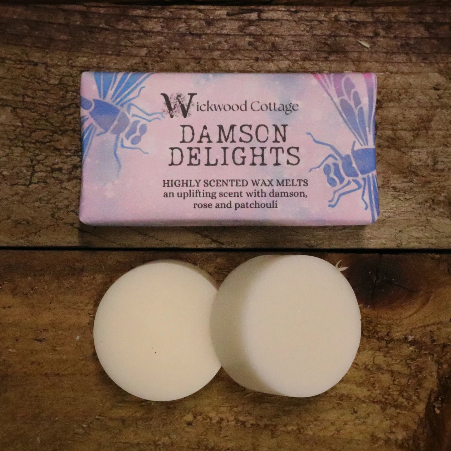 A rectangle paper package of 'Damson Delights' wax melts by Wickwood Cottage, featuring highly scented notes of damson, rose, and patchouli. The packaging is pink with dragon fly patterns. Below the package is an example of what's inside, two 20g wax melt discs and the product is displayed on a rustic wooden table.