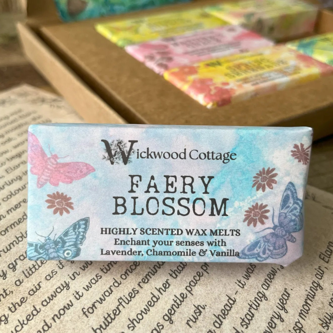 Paper packaging of Wickwood Cottage Faery Blossom highly scented wax melts. The packaging has a butterfly pattern with blue background and informative text. In the background is the gift box. Underneath is the story which comes with the gift box.