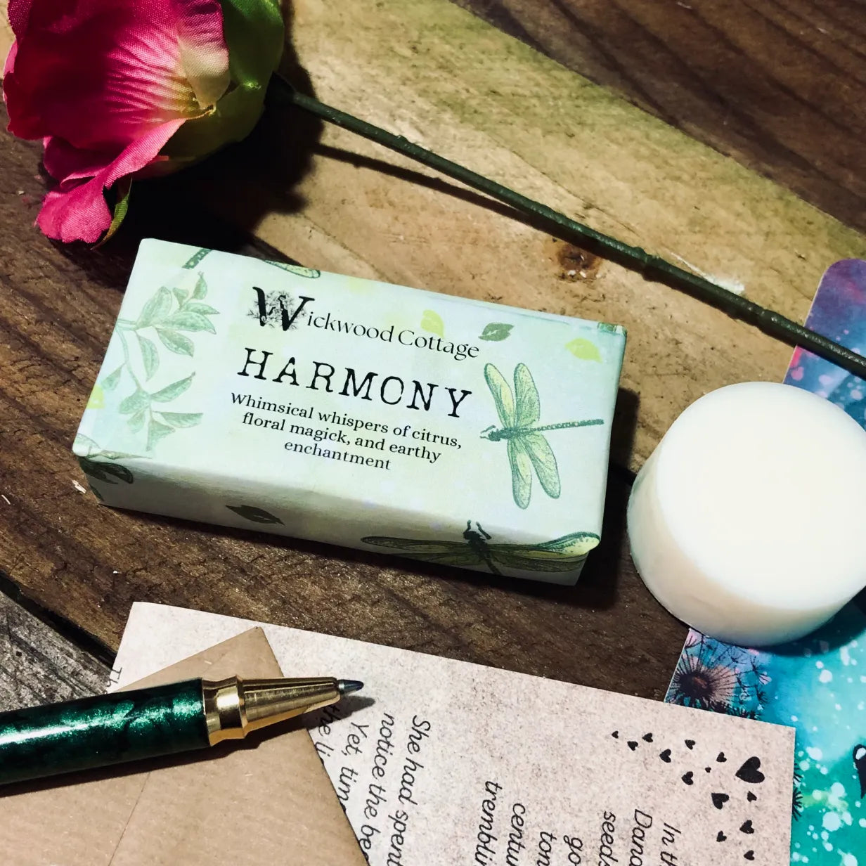 Paper packaging of Wickwood Cottage Harmony highly scented wax melts. The packaging has dragonfly's and a floral pattern with green background. The image is set within a rustic table depicting a loved one's letter on the table a single rose a pen and the story and postcard.