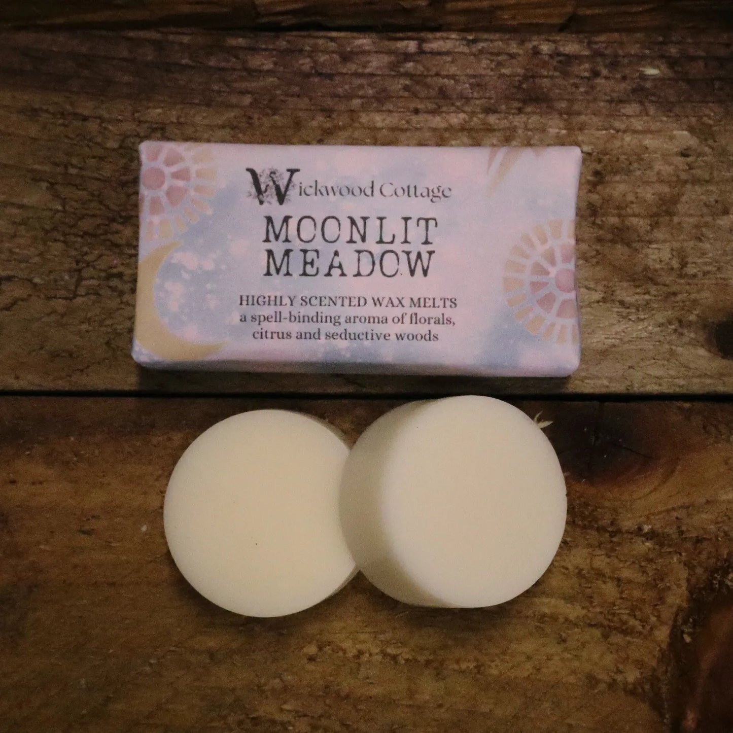 A rectangle paper package of 'Moonlit Meadow' wax melts by Wickwood Cottage. The packaging is lilac with a floral pattern and descriptive text, displayed on top of a rustic wooden table. Below is an example of its contents, two wax melt discs