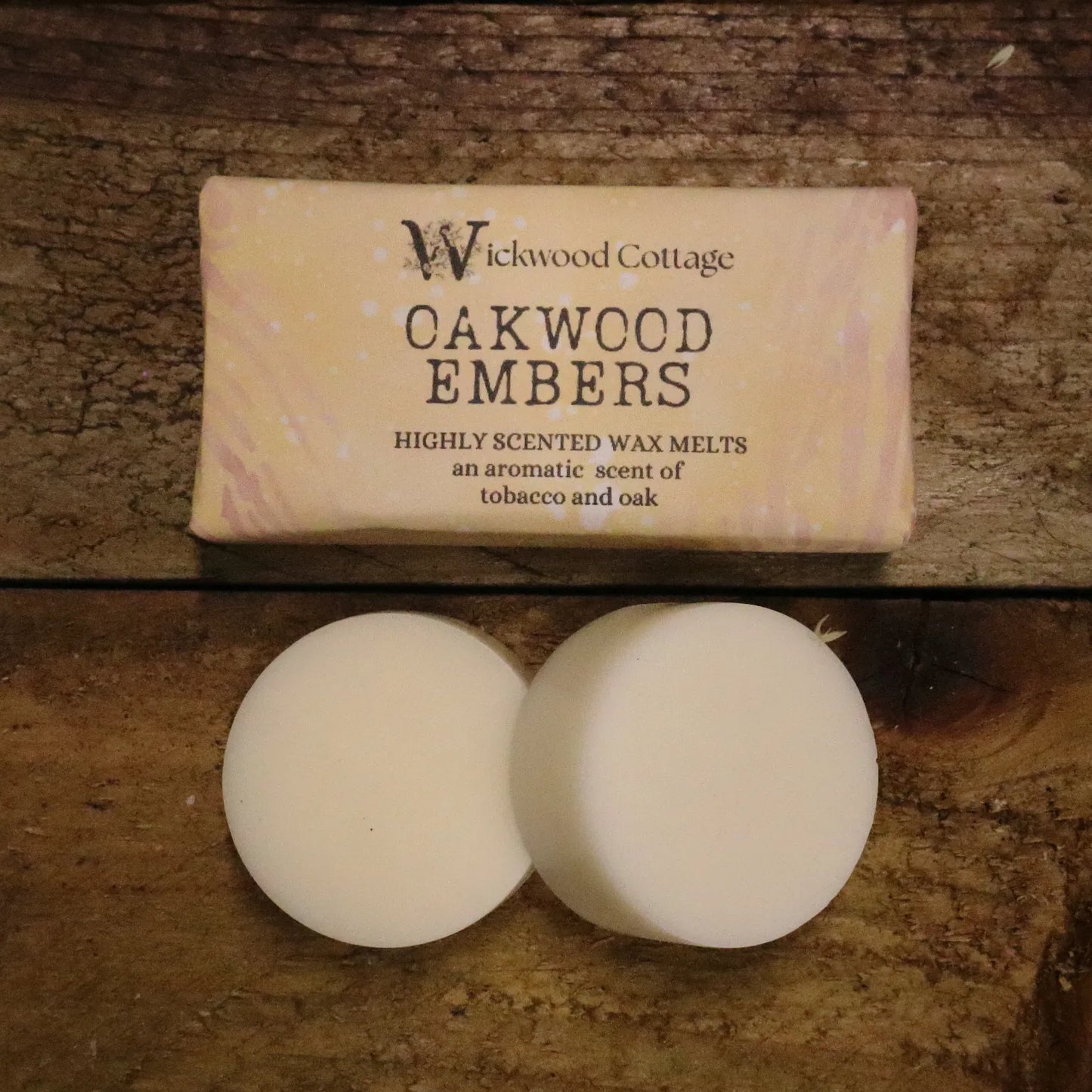 A rectangle paper package of 'Oakwood Embers' wax melts by Wickwood Cottage. The packaging is light brown with a tree bark pattern and descriptive text, displayed on top of a rustic wooden table. Below the package are two wax melt discs showing the contents.