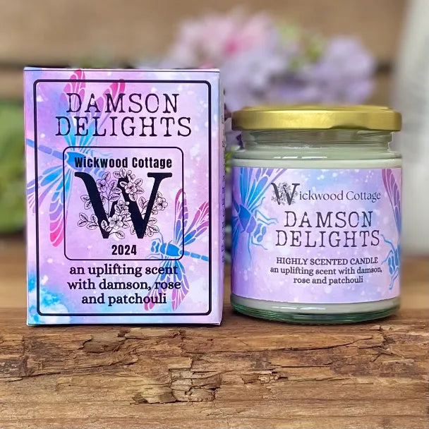 Wickwood Cottage Damson Delights candle and packaging on a rustic wooden surface with a blurred floral background. To the left is the candle box pale violet with blue dragonflies and black text. To the right is the candle a 150-gram candle jar with a gold lid.