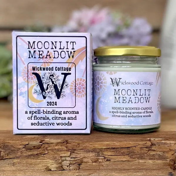 Wickwood Cottage Moonlit Meadow candle and packaging on a rustic wooden surface with a blurred floral background. To the left is the candle box pale blue with gold florals and black text. To the right is the candle a 150-gram candle jar with a gold lid.