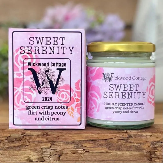 Wickwood Cottage Sweet Serenity candle and packaging on a rustic wooden surface with a blurred floral background. To the left is the candle box pink with darker pink florals and black text. To the right is the candle a 150-gram candle jar with a gold lid.