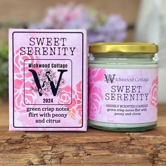 Wickwood Cottage Sweet Serenity candle and packaging on a rustic wooden surface with a blurred floral background. To the left is the candle box pink with darker pink florals and black text. To the right is the candle a 150-gram candle jar with a gold lid.
