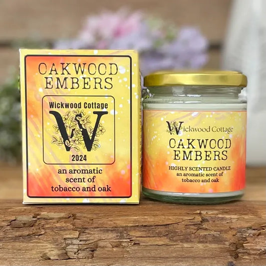 Wickwood Cottage Oakwood Embers candle and packaging on a rustic wooden surface with a blurred floral background. To the left is the candle box yellow and red with black text. To the right is the candle a 150-gram candle jar with a gold lid.