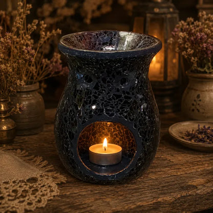 A black crackle-glaze ceramic oil burner candle holder with a tea light candle inside, set on a rustic wooden table with dried flowers and warm ambient background lighting.