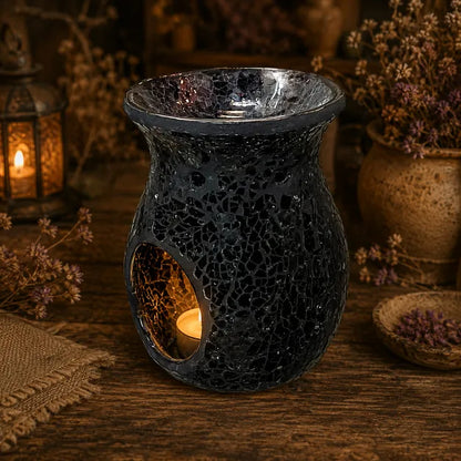 A dark crackle-glazed ceramic candle warmer with a small tea light candle glowing inside, styled on a rustic wooden surface with dried flowers and a lantern in the background.