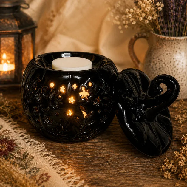 A black ceramic pumpkin-shaped wax warmer with carved floral details and a matching lid leaning beside it, set on a wooden surface with warm candlelight glowing through the cutouts. A lit lantern, draped fabric, and dried flowers appear in the softly blurred background.