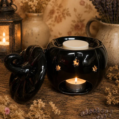 A glossy black pumpkin-shaped wax warmer with a matching lid, shown on a rustic wooden surface with dried flowers and a lantern in the background. The warmer contains a white wax melt on top and a tea light candle inside, with small cutout details showing warm light.