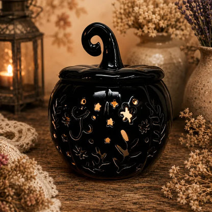 A black ceramic Halloween pumpkin wax warmer with a pointed lid and cutout details, shown on a rustic wooden surface surrounded by dried flowers and a warm lantern in the background.