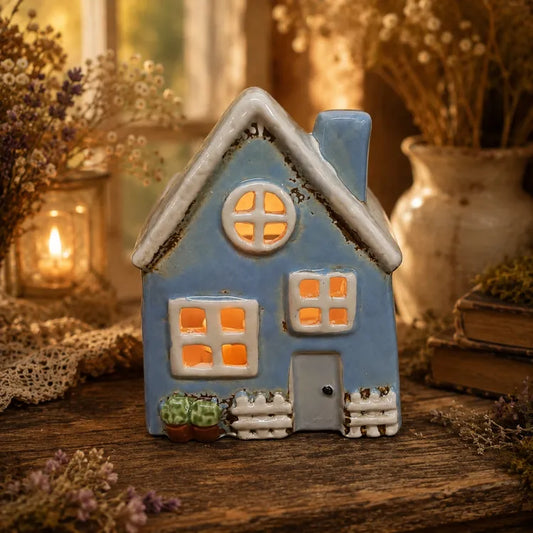 A small blue ceramic house figurine with a white roof, glowing orange windows, and a grey door, displayed on a rustic wooden surface with dried flowers and warm candlelight in the background.