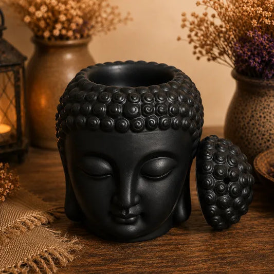 A black Buddha head wax/oil warmer with its lid resting beside it on a wooden surface, styled with dried flowers and a lantern in the background.