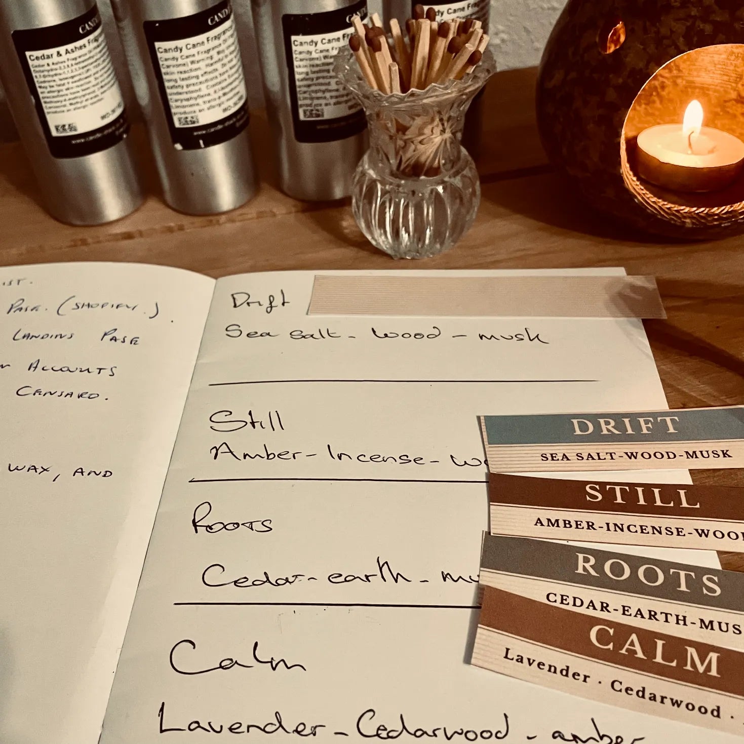 Censaro scent planning scene with handwritten fragrance notes in a notebook, printed labels for Drift, Still, Roots and Calm, fragrance oil bottles, matches and a glowing candle burner in the background.
