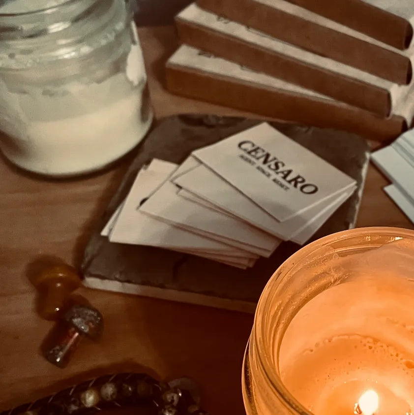 Censaro work-in-progress scene with printed brand labels on a slate tile, brown packaging materials, wax supplies and a glowing candle jar in warm ambient light.