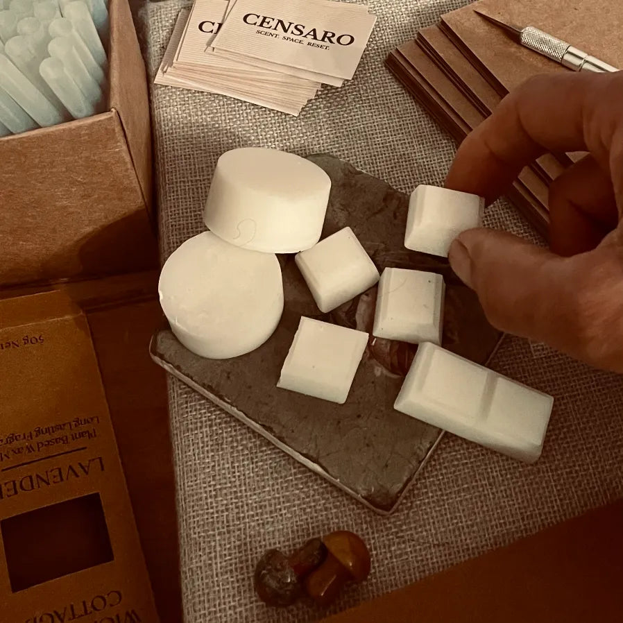 Censaro home fragrance work-in-progress scene showing white wax melt samples on a slate tile, brown packaging materials, printed Censaro labels and a hand selecting a wax piece.