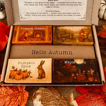 An open box of Autumn wax melts by Wickwood Cottage the Cosy Nights Collection. Four wax melts with different fragrances and packaging. Sitting on a rustic wooden surface and surrounded by autumn leaves, pumpkins, toadstools and two white wax melt discs. 