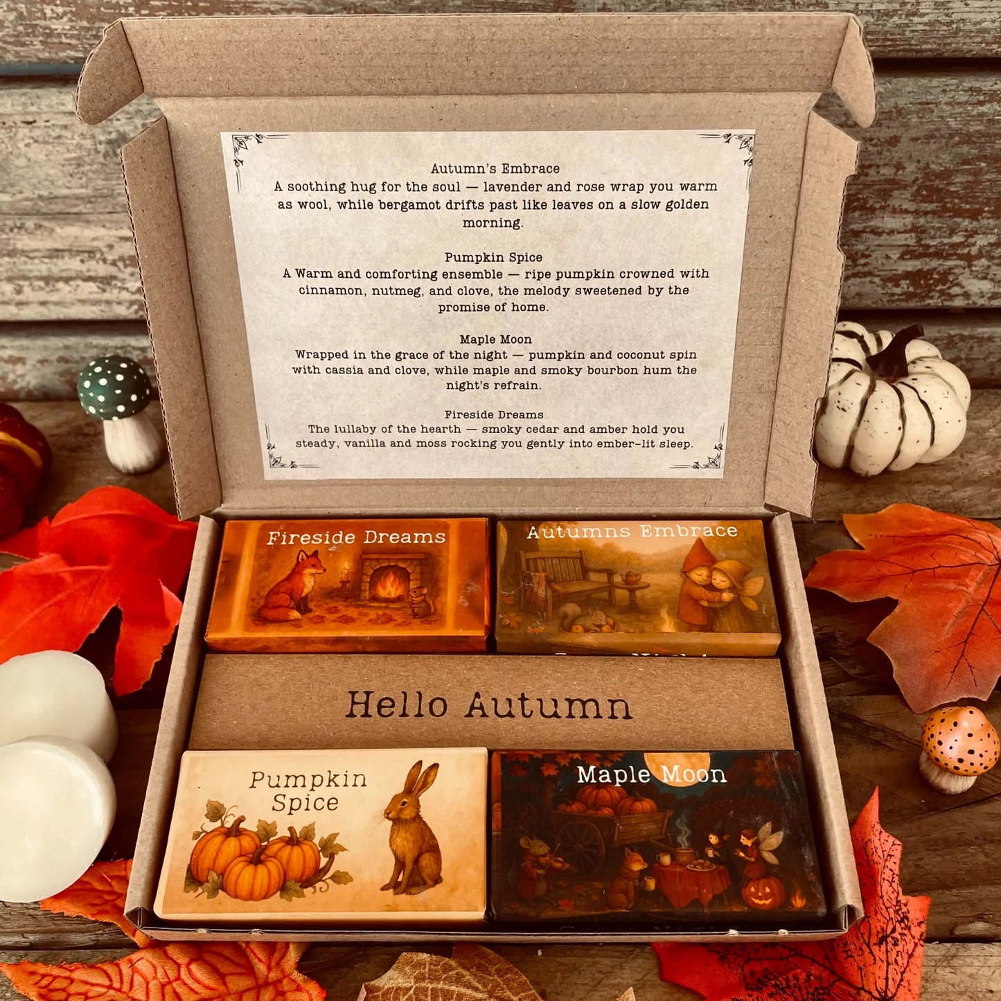 An open box of Autumn wax melts by Wickwood Cottage the Cosy Nights Collection. Four wax melts with different fragrances and packaging. Sitting on a rustic wooden surface and surrounded by autumn leaves, pumpkins, toadstools and two white wax melt discs. 