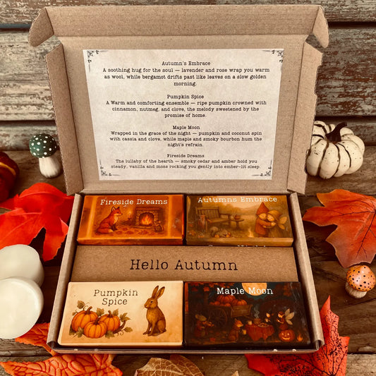 An open box of Autumn wax melts by Wickwood Cottage the Cosy Nights Collection. Four wax melts with different fragrances and packaging. Sitting on a rustic wooden surface and surrounded by autumn leaves, pumpkins, toadstools and two white wax melt discs. 