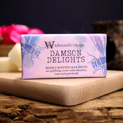 A rectangle paper package of 'Damson Delights' wax melts by Wickwood Cottage, featuring highly scented notes of damson, rose, and patchouli. The packaging is pink with dragon fly patterns and the product is displayed on a wooden platter on top of a rustic wooden table. 