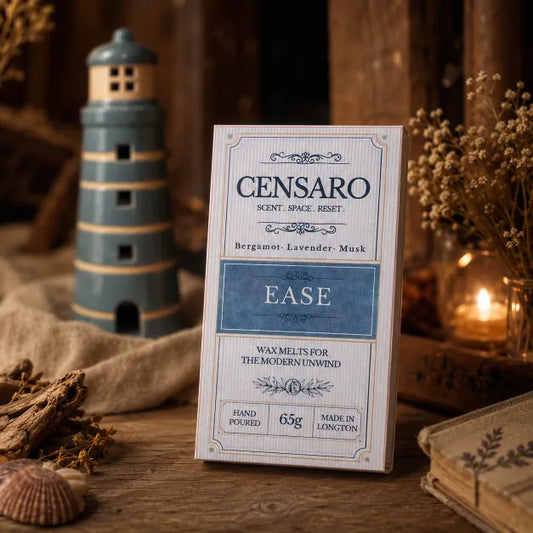 A boxed fragrance product labeled CENSARO EASE stands upright on a rustic wooden surface with dried flowers, a lighthouse decoration, and a warm candle in the background. The box is cream and blue, and the scene is styled in a cozy indoor setting.