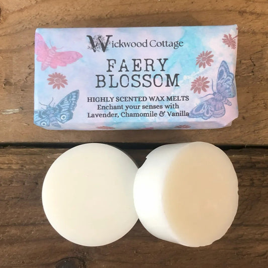 Two white wax melt discs displayed on a rustic wooden surface beneath a Wickwood Cottage label reading “Faery Blossom,” decorated with butterflies and soft pastel floral artwork.