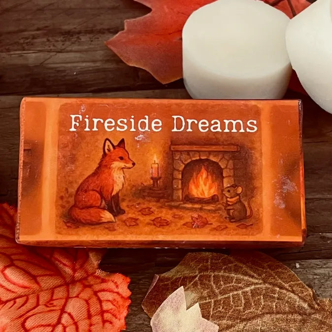 Paper wrapped rectangle wax melt with 'Fireside Dreams' text and an image of a fox and mouse sitting Infront of a roaring coal fire. Surrounded by autumn leaves a pumpkin and two wax melt discs on a rustic wooden surface. This is a close-up image.