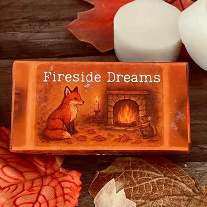 Paper wrapped rectangle wax melt with 'Fireside Dreams' text and an image of a fox and mouse sitting Infront of a roaring coal fire. Surrounded by autumn leaves a pumpkin and two wax melt discs on a rustic wooden surface. This is a close-up image.