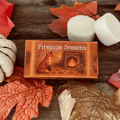 Paper wrapped rectangle wax melt with 'Fireside Dreams' text and an image of a fox and mouse sitting Infront of a roaring coal fire. Surrounded by autumn leaves a pumpkin and two wax melt discs on a rustic wooden surface.