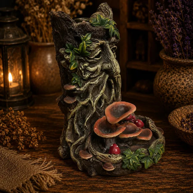 A decorative tree stump figurine with a carved forest face, green leaves, and layered brown mushrooms with small red toadstools, displayed on a wooden surface in a rustic styled setting with dried plants and a lantern.