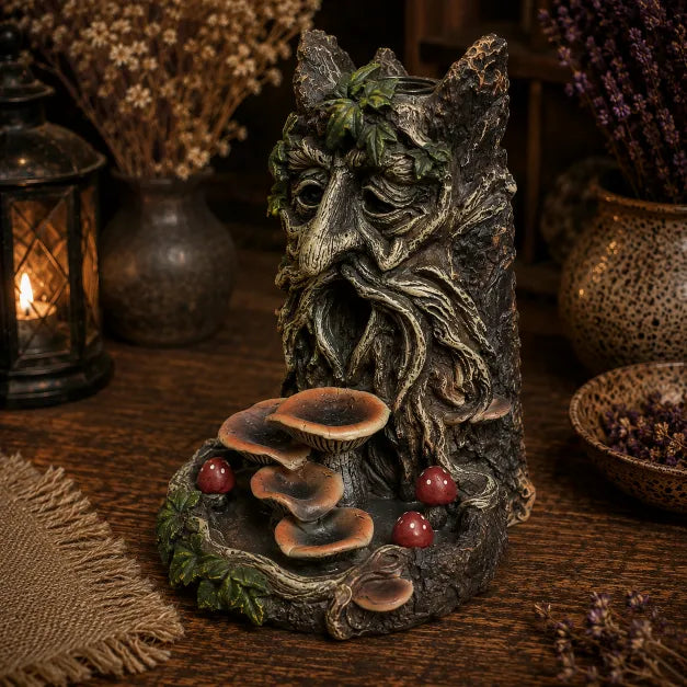 A decorative fantasy tree-man backflow sculpture with a carved forest face, leafy crown, mushrooms, and small red toadstools on a rustic wooden surface, surrounded by dried flowers and a lantern in the background.