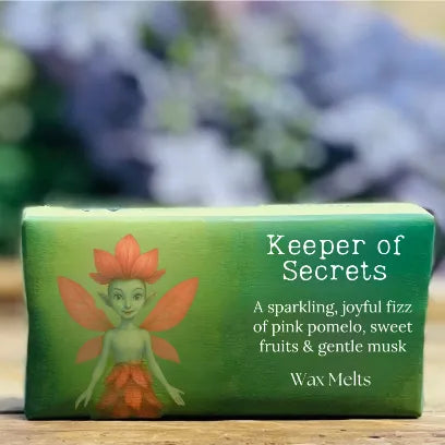 A rectangular pack of handmade strong-smelling wax melts featuring Petalune, a green faery with pink petal wings and a flower crown. She stands smiling on the left side of the packaging, while the right side displays the scent description for the Keeper of Secrets fragrance. The box rests on a rustic wooden surface.