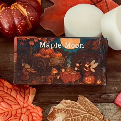 Paper wrapped rectangle wax melt with 'Maple Moon' text and an image of an enchanted garden party with a faery and dressed woodland creatures. Surrounding the package, autumn leaves a pumpkin and two wax melt discs on a rustic wooden surface. This is a close up image
