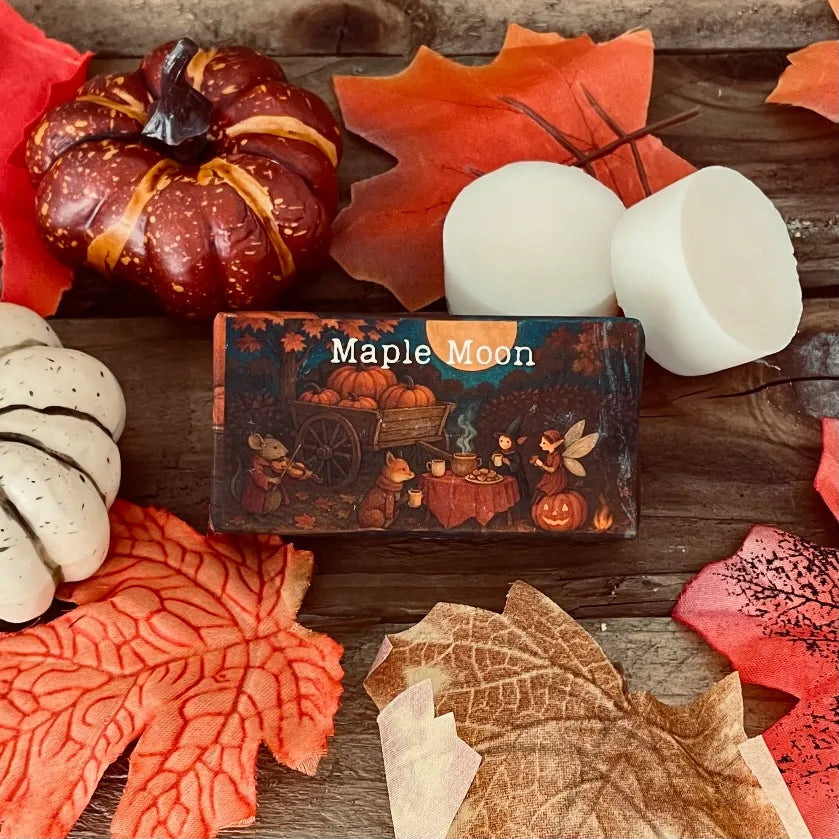 Paper wrapped rectangle wax melt with 'Maple Moon' text and an image of an enchanted garden party with a faery and dressed woodland creatures. Surrounding the package, autumn leaves a pumpkin and two wax melt discs on a rustic wooden surface.