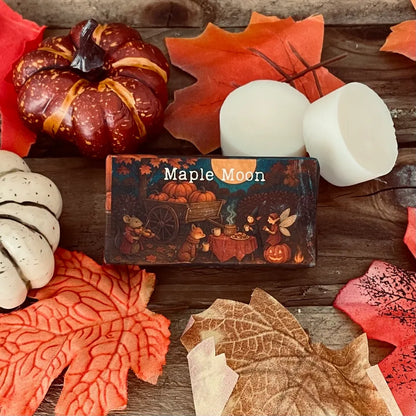 Paper wrapped rectangle wax melt with 'Maple Moon' text and an image of an enchanted garden party with a faery and dressed woodland creatures. Surrounding the package, autumn leaves a pumpkin and two wax melt discs on a rustic wooden surface.
