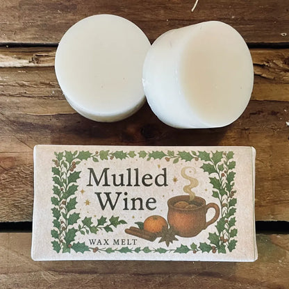 Two round Mulled Wine wax melts resting beside their festive woodland-themed label featuring a steaming mug, orange slice and spices, displayed on a rustic wooden surface; strong smelling wax melts from Wickwood Cottage.