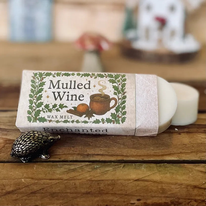 Mulled Wine wax melts wrapped in a festive woodland label with an illustration of a steaming mug, orange slice and spices, displayed on a rustic wooden surface with a small hedgehog figurine; strong smelling wax melts from Wickwood Cottage.