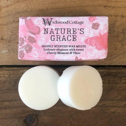 Two white wax melt discs displayed on ra ustic wooden surface beneath a Wickwood Cottage label reading “Nature’s Grace,” decorated with cherry blossom and butterfly artwork.