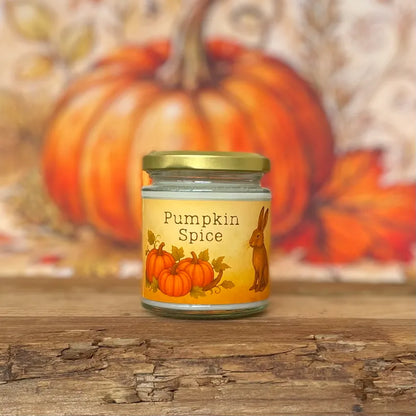 Pumpkin spice fragranced candle in a jar with pumpkins and a hair on the label and a gold lid, set against a blurred pumpkin background on a rustic wooden table,