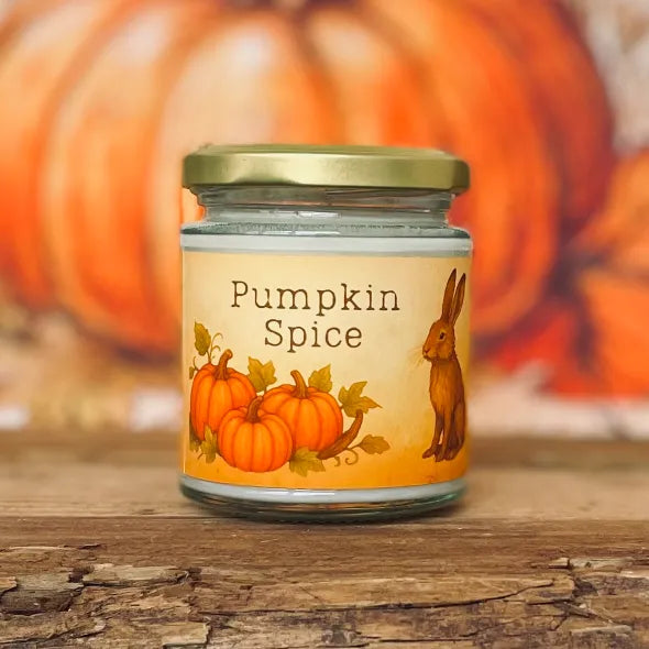 Pumpkin spice fragranced candle in a jar with pumpkins and a hair on the label and a gold lid, set against a blurred pumpkin background on a rustic wooden table.