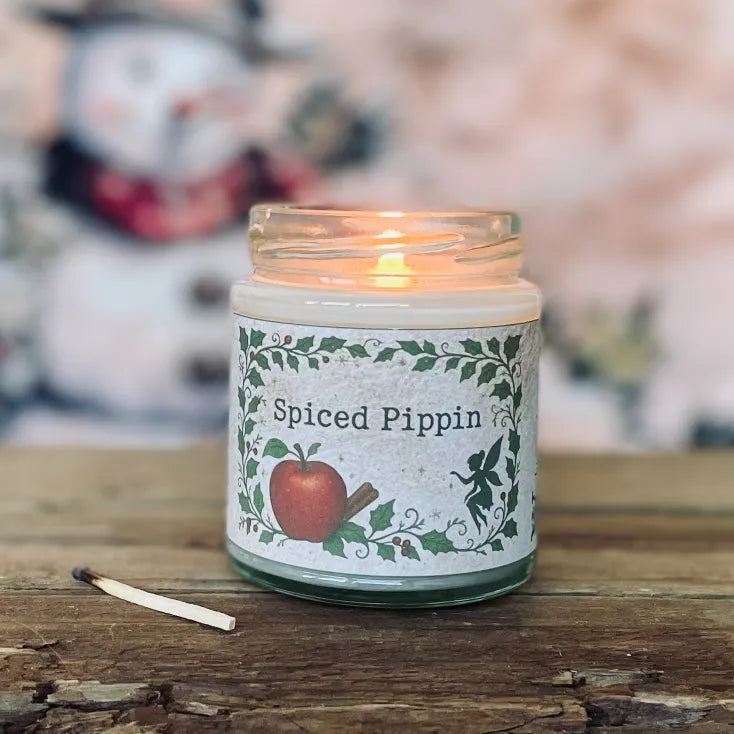 Lit Spiced Pippin fragranced candle in a glass jar with woodland apple and cinnamon label, sitting on a rustic wooden surface with a used match; a cosy magickal candle from Wickwood Cottage.