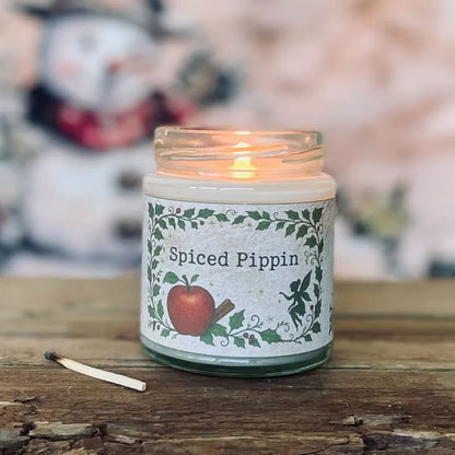 Lit Spiced Pippin fragranced candle in a glass jar with woodland apple and cinnamon label, sitting on a rustic wooden surface with a used match; a cosy magickal candle from Wickwood Cottage.