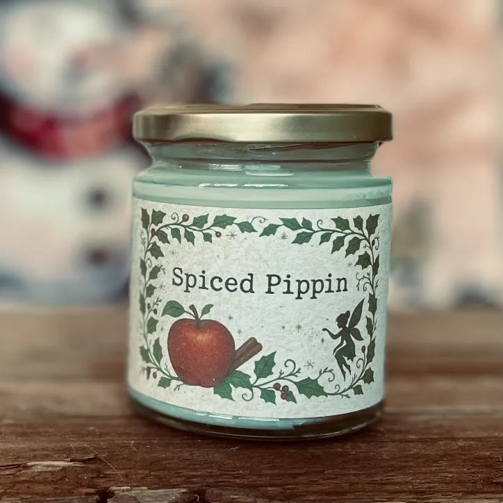 Spiced Pippin 150g fragranced candle in a glass jar with gold lid and woodland label featuring an apple, cinnamon stick, holly and fairy design; a cosy magickal candle from Wickwood Cottage.