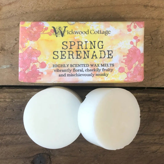 Two white wax melt discs displayed on a rustic wooden surface beneath a Wickwood Cottage label reading “Spring Serenade,” decorated with vibrant floral artwork in warm spring colours